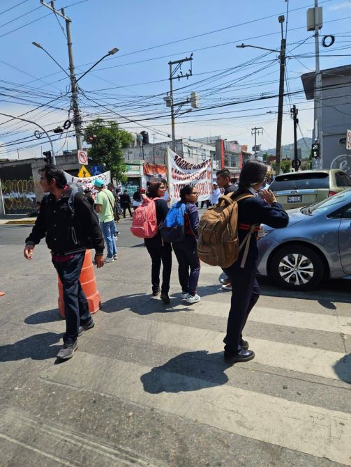 Grupo de personas cruzando la calle, una manifestación visible al fondo, con carteles y tráfico cercano.