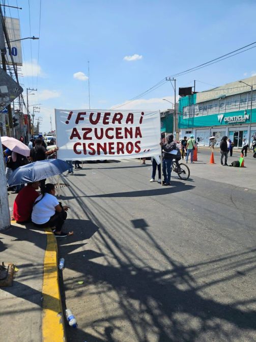 Manifestación en la calle con una pancarta que dice '¡Fuera! Azucena Cisneros'. Varias personas están presentes, algunas bajo sombrillas.