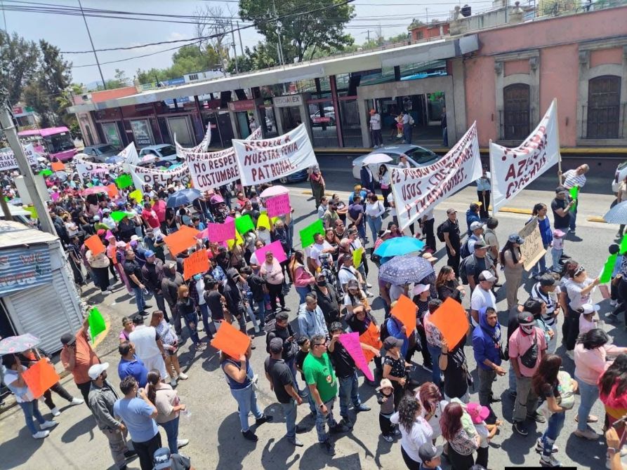 Protesta con numerosos participantes sosteniendo pancartas y carteles de colores brillantes en una calle, junto a un transporte público.