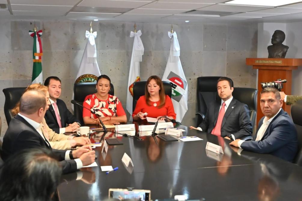 Reunión de varias personas en una mesa de conferencia, con banderas y un busto en el fondo.