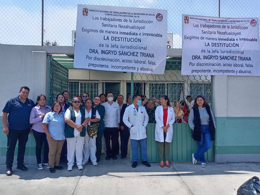 Grupo de trabajadores de la Jurisdicción Sanitaria Nezahualcóyotl frente a un edificio, sosteniendo carteles que exigen la destitución de la Dra. Ingryd Sánchez Triana por razones laborales.