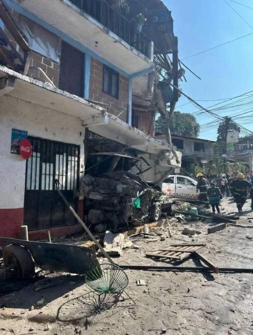 Escena de destrucción tras un accidente, mostrando un edificio dañado, un automóvil impactado y escombros en la calle. Hay equipos de emergencia trabajando en el lugar.