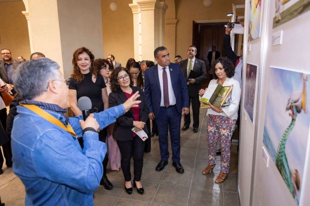 Un grupo de personas en una galería de arte, observando obras mientras un hombre habla con un micrófono.