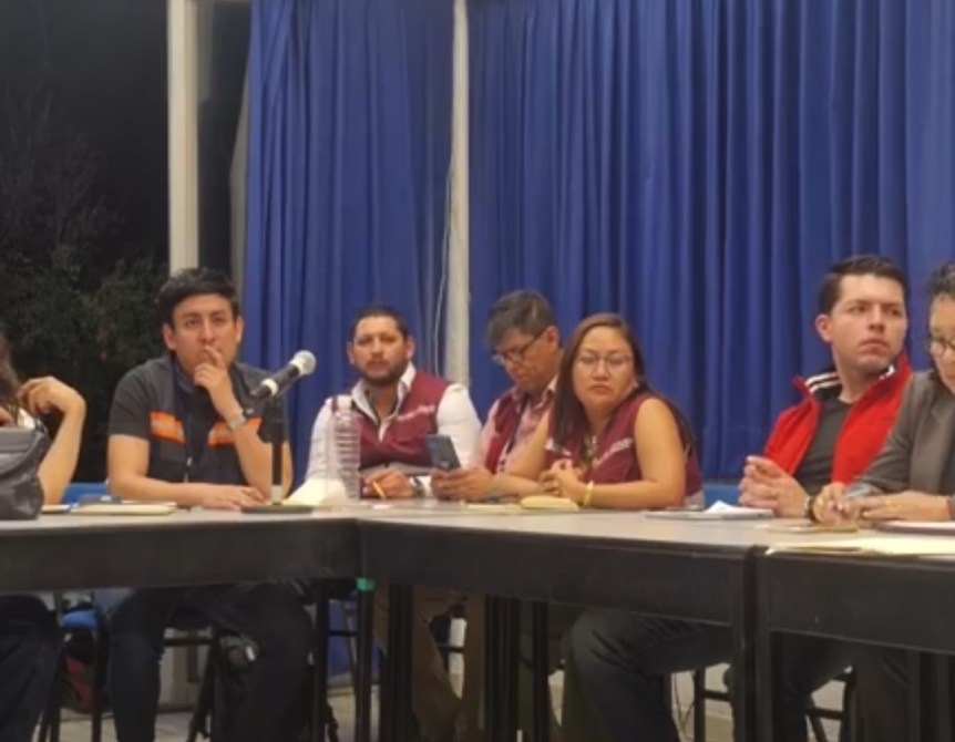 Un grupo de personas sentadas alrededor de una mesa en una reunión, con fondo de cortinas azules.