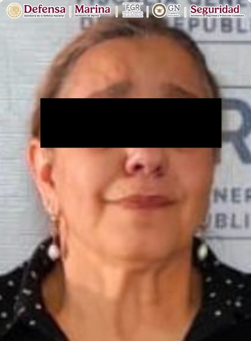 Mujer con cabello castaño claro, mirando hacia adelante, con expresión neutral. Lleva pendientes y una blusa negra con puntos blancos. Fondo azul claro.