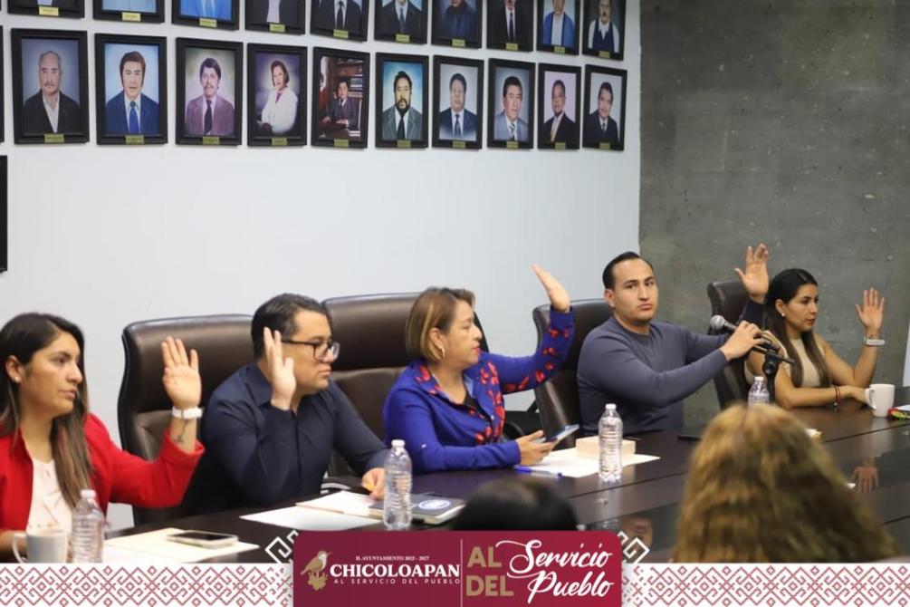 Reunión de funcionarios levantando la mano en señal de aprobación, con retratos en la pared de fondo.