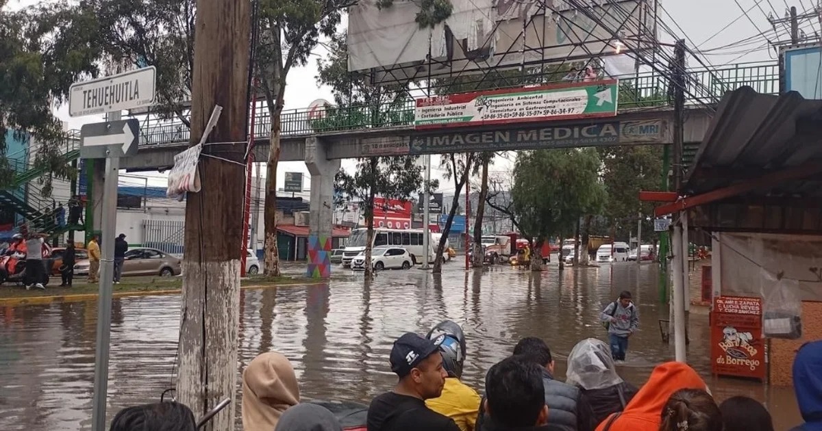 Inundación en una calle con vehículos parcialmente sumergidos y personas tratando de cruzar el agua, señal de tránsito visible en el lado izquierdo.