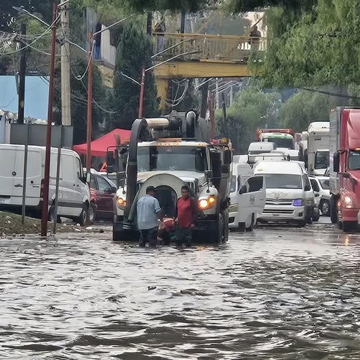 Inundación en una calle con vehículos atrapados, personas en el agua y un camión de carga en el centro de la imagen.