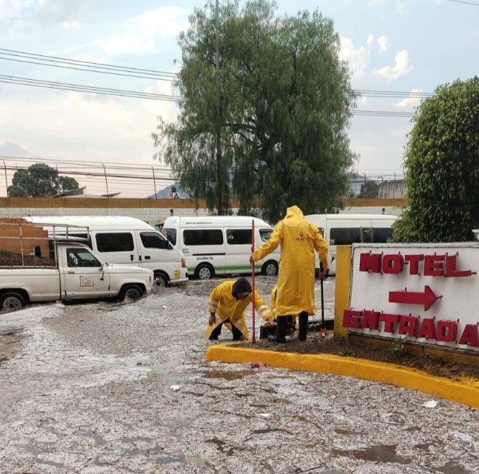 Dos trabajadores en trajes de lluvia amarillos limpiando agua y escombros en la entrada de un motel, rodeados de vehículos y un árbol.