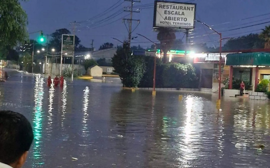Inundación en una calle con personas caminando, de noche, con señalización de un restaurante y un hotel en el fondo.