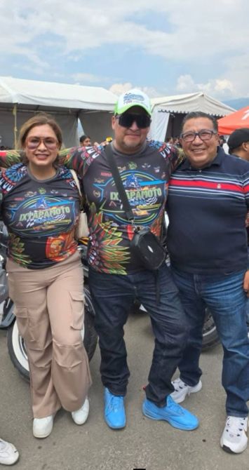 Tres personas posando juntas en un evento, vistiendo camisetas coloridas con un diseño relacionado con motocicletas. Una persona lleva una gorra y gafas de sol, mientras que las otras dos sonríen. Al fondo, se pueden ver carpas y un ambiente festivo.