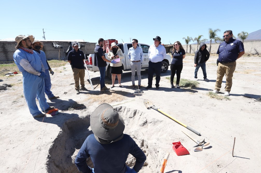 A group of workers and officials observing an excavation site, with one person digging, while others are engaged in discussion nearby.