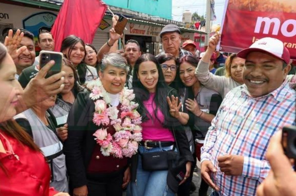 Grupo de personas sonrientes en un evento al aire libre, algunas llevan banderas y otras hacen fotos con sus teléfonos. Hay una mujer con un collar de flores en el centro de la imagen.
