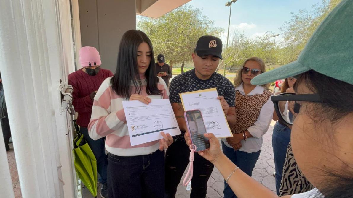 Dos personas sosteniendo documentos frente a un grupo, mientras otra persona graba con un teléfono móvil.