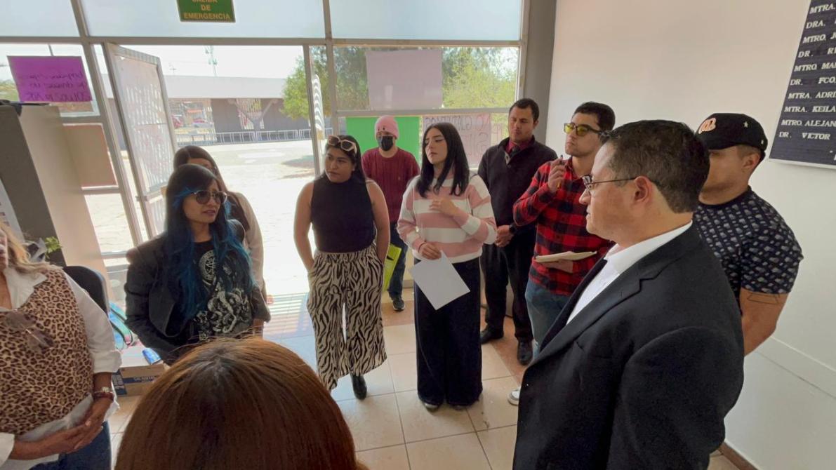 Grupo de personas reunidas en un espacio interior, algunas de pie en círculo mientras escuchan a un orador. Hay una ventana grande al fondo y documentos en manos de algunos asistentes.