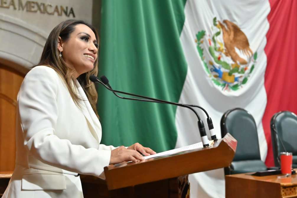 Mujer hablando en un podio dentro de un recinto legislativo, con banderas de México de fondo.