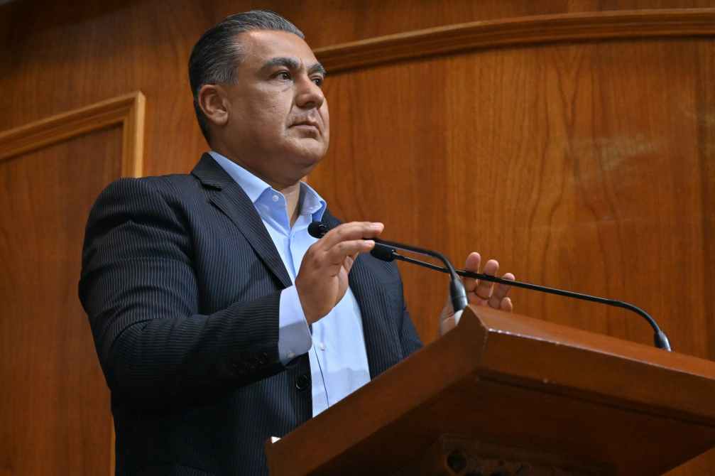 Hombre de pie frente a un atril, hablando con las manos gesticulando, en un entorno de oficina con paneles de madera.