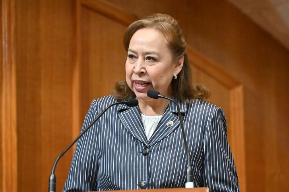 Mujer de pie hablando en un micrófono en un escenario, con un fondo de madera.