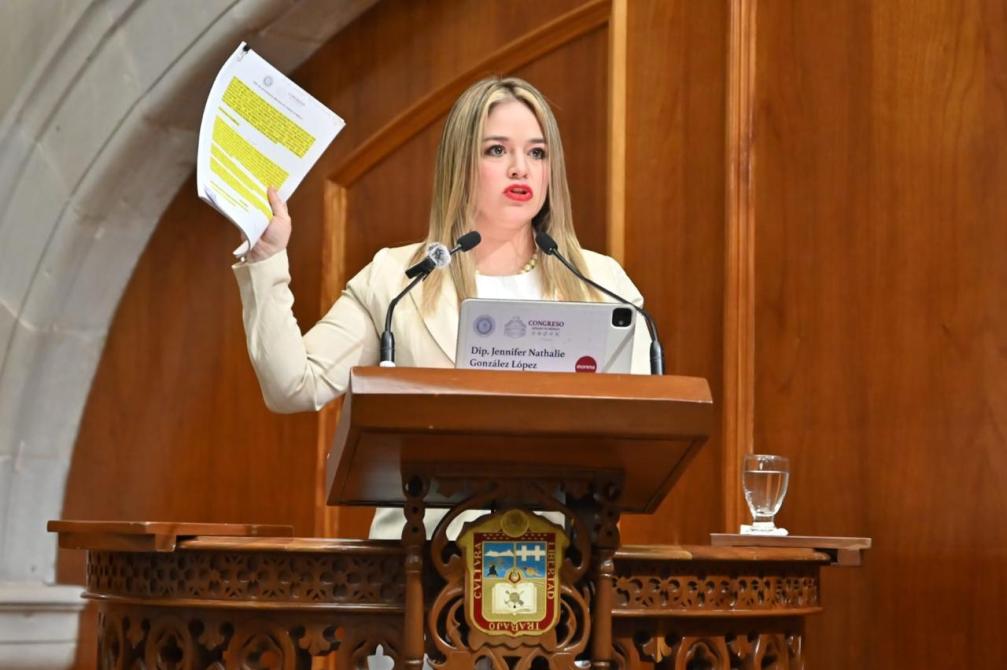 Una diputada de pie en un podio, sosteniendo documentos con marcas resaltadas, mientras habla en una sesión del congreso.