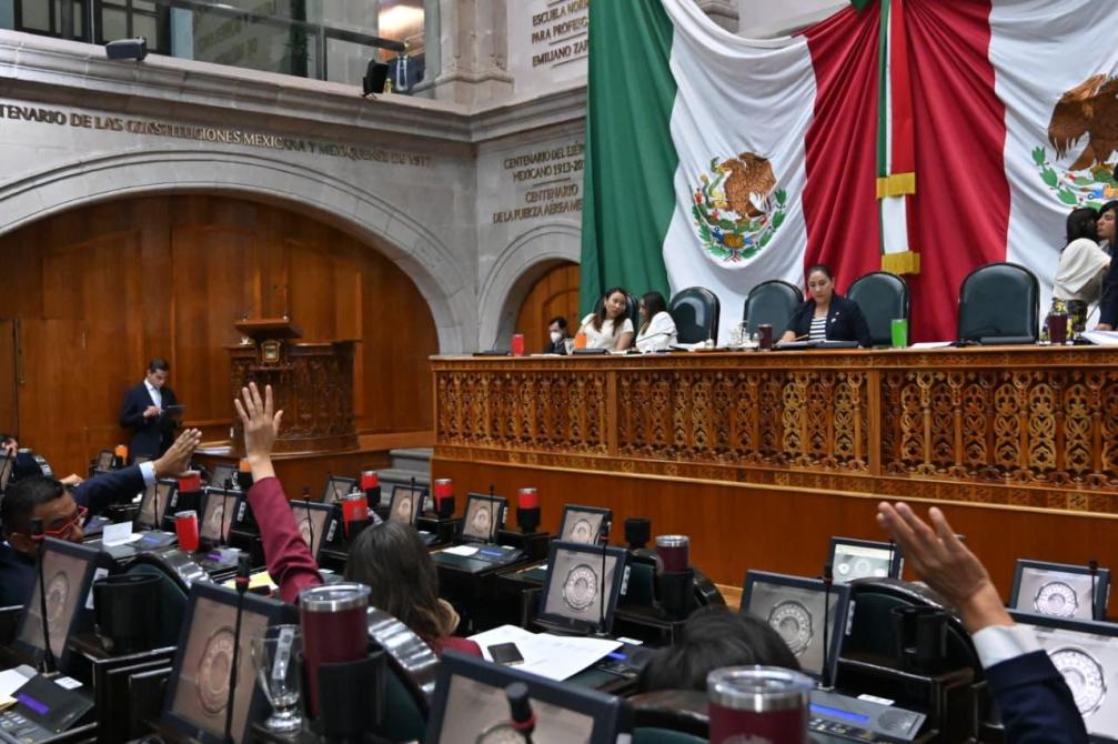 Sesión legislativa en un recinto oficial con representantes levantando la mano para votar, banderas de México en el fondo.