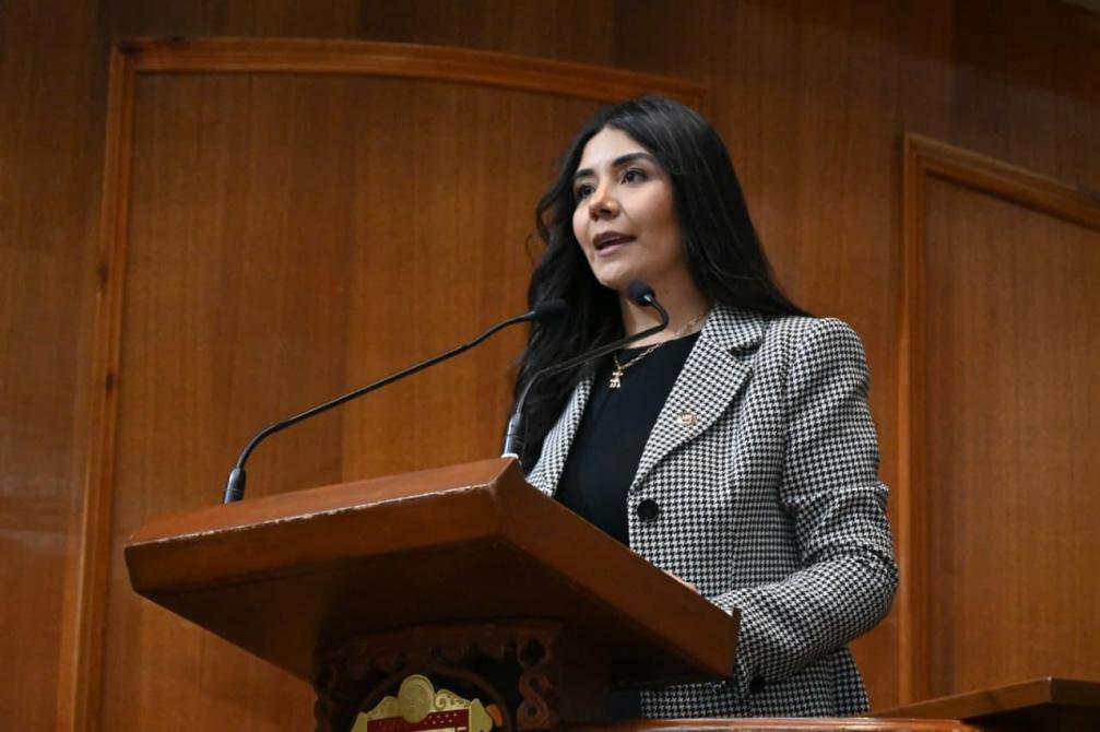 Una mujer habla desde un podio en un ambiente formal, con un fondo de madera y luces suaves.