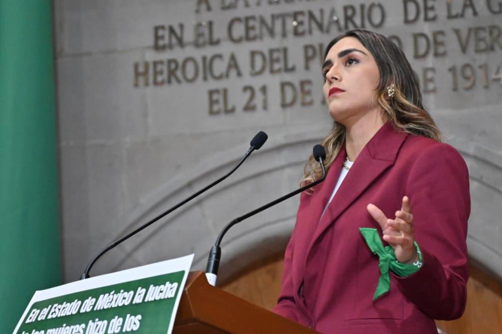 Mujer de pie en un podio hablando, con una chaqueta color burdeos y un moño verde. Fondo con una pared que tiene inscripciones históricas.