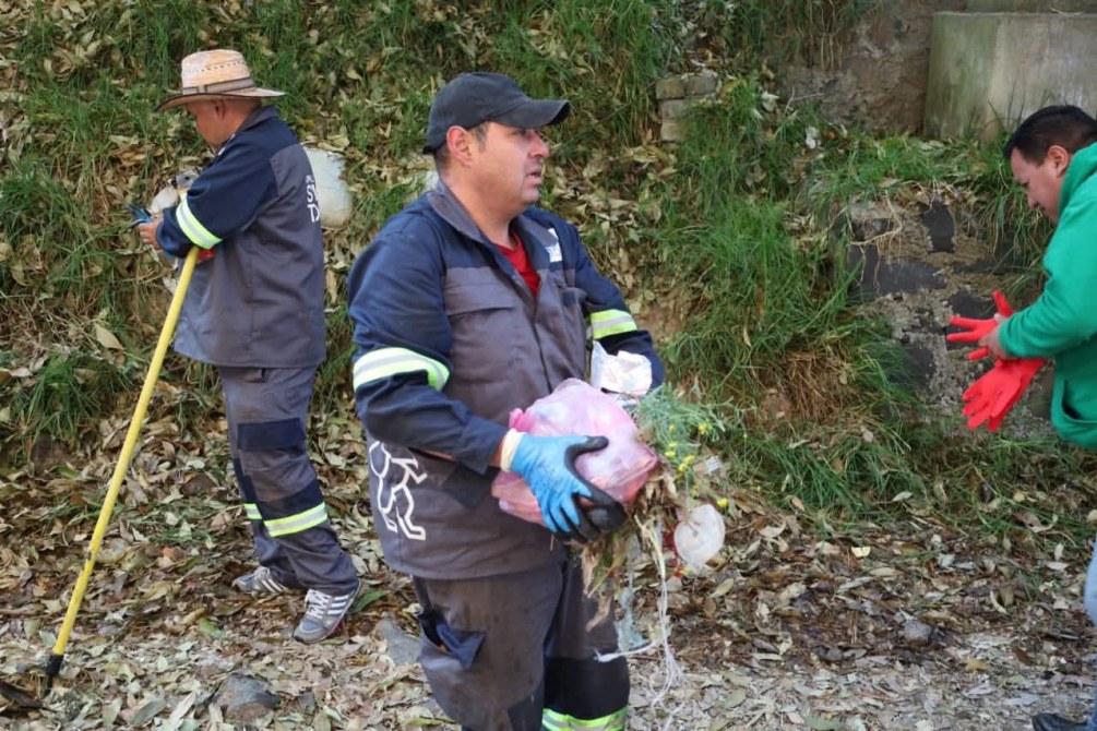 Trabajadores de limpieza en un área verde, recogiendo desechos y hojas secas. Uno lleva guantes y sostiene un bulto, mientras otro usa una pala.