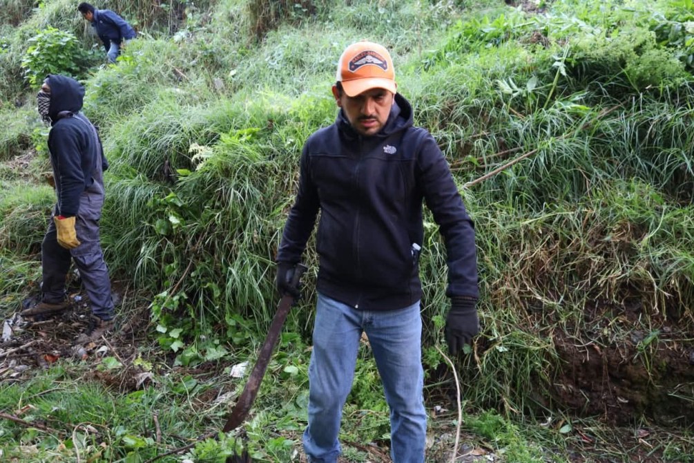 Grupo de personas trabajando en un área verde, un hombre con gorra naranja y guantes sostiene una herramienta en un paisaje de hierba.