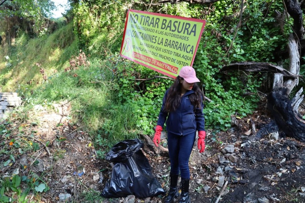 Una persona con guantes rojos y ropa de trabajo, de pie junto a un aviso que dice 'NO TIRAR BASURA', en un área verde con desechos en el suelo.