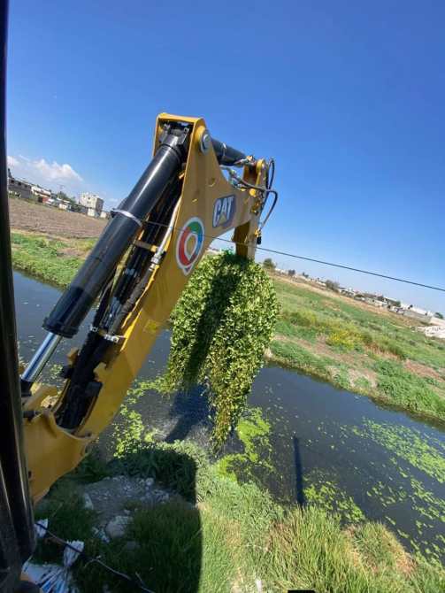 Excavadora amarilla CAT levantando vegetación acuática sobre un canal.