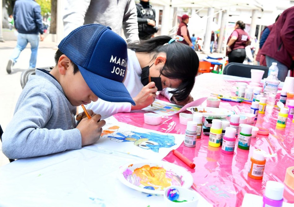 Niño y niña pintando en una mesa con pinturas y materiales de arte, en un evento al aire libre.