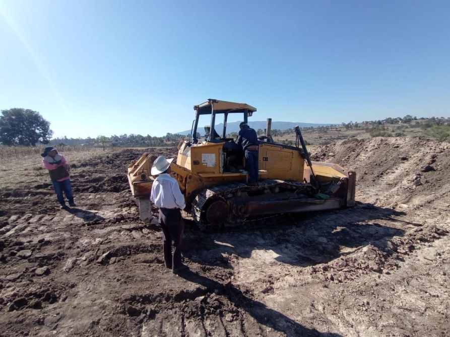 A yellow bulldozer operating on a construction site, with two workers observing and one worker on the machine. The landscape shows cleared earth and a clear blue sky.