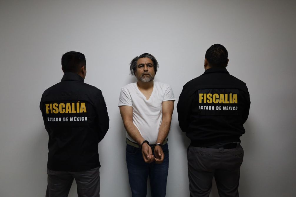 Hombre en camiseta blanca, esposado y de pie frente a una pared, flanqueado por dos oficiales de la Fiscalía del Estado de México, que llevan chaquetas negras con el logo visible.