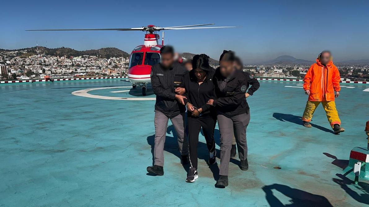 Una persona detenida siendo escoltada por dos agentes de seguridad en una azotea, con un helicóptero de fondo y una vista de la ciudad.