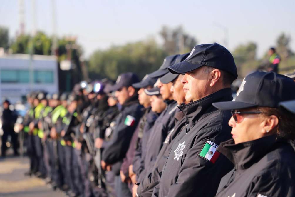 Grupo de policías en formación, con uniformes negros y gorras, destacando una bandera mexicana en algunas prendas.