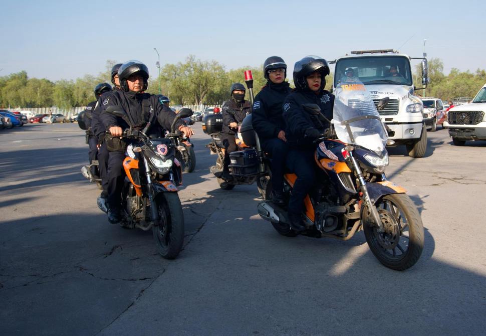 Grupo de motociclistas policías en una carretera, algunos llevan cascos y uniformes oscuros, mientras que se observa tráfico de vehículos detrás.