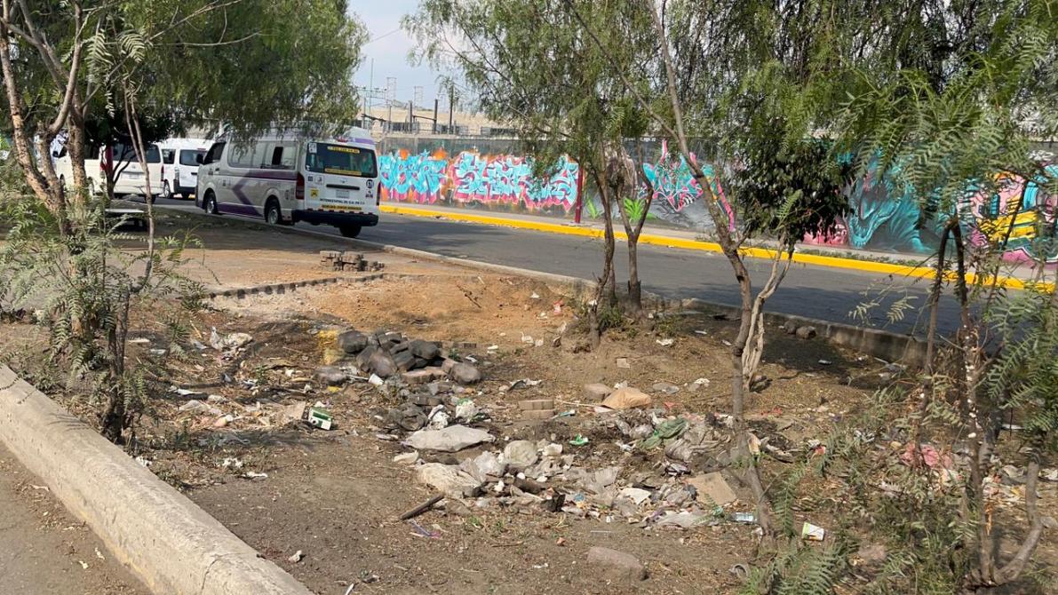 Vista de una zona desordenada con basura y maleza, junto a una carretera y un mural colorido al fondo.