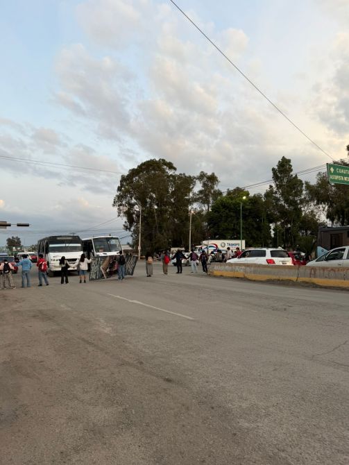 Grupo de personas bloqueando una carretera, con autobuses y vehículos estacionados.