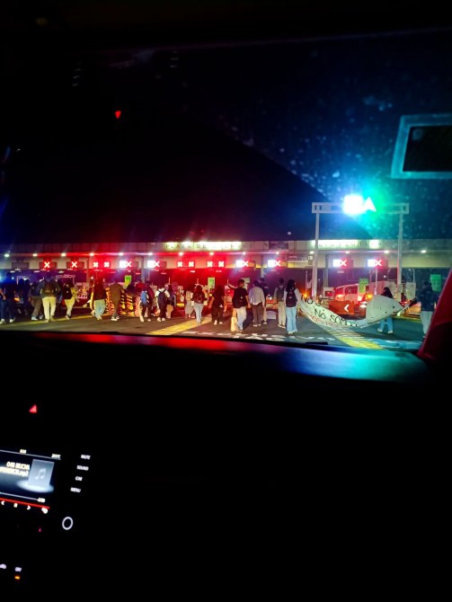Grupo de personas en la calle durante la noche, con luces rojas y una señal verde visible, frente a un edificio.