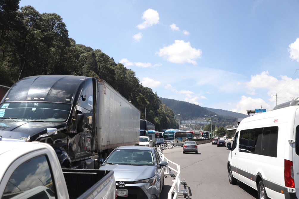 Tráfico vehicular en una carretera con camiones y autos, rodeado de árboles y montañas bajo un cielo azul con nubes blancas.