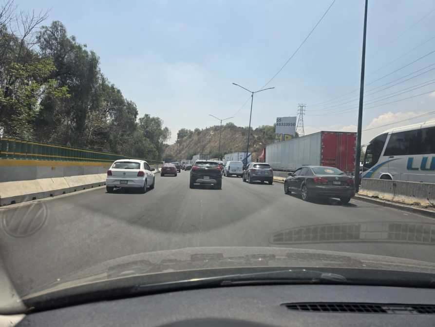 Vista de una carretera con varios automóviles en movimiento, incluyendo un camión grande y un autobús, rodeada de árboles y un cerro al fondo.