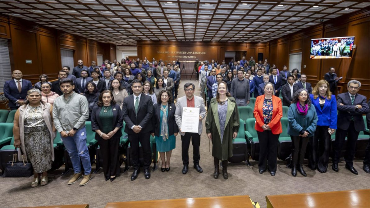 Grupo de personas posando en una sala de conferencias, algunas sosteniendo un documento, con un fondo de sillas verdes y pantallas de presentación.