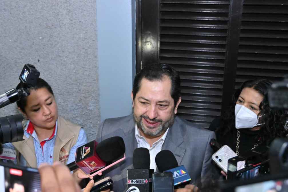 Un hombre con barba y traje, rodeado de varios micrófonos y periodistas, sonríe mientras habla, mientras dos mujeres lo escuchan atentamente.