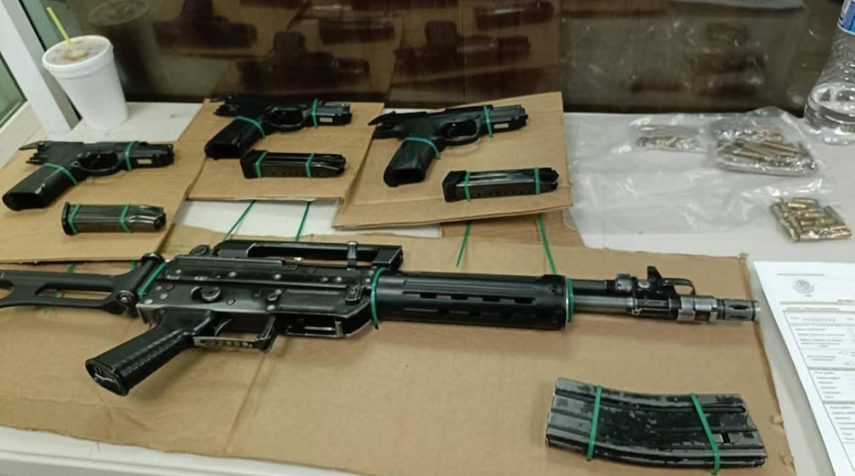 Armas de fuego y municiones dispuestas sobre cartón, incluyendo un fusil y varias pistolas.