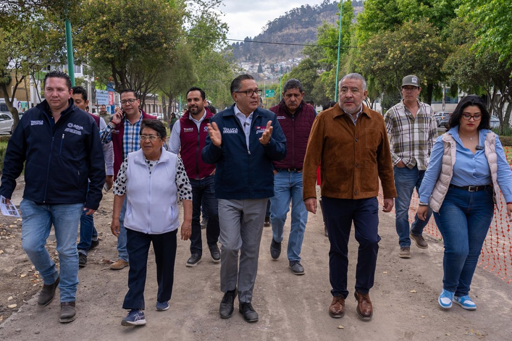 Un grupo de personas caminando por una calle en un ambiente urbano; algunos llevan ropa de trabajo y otros casual. Se observa vegetación y montañas al fondo.