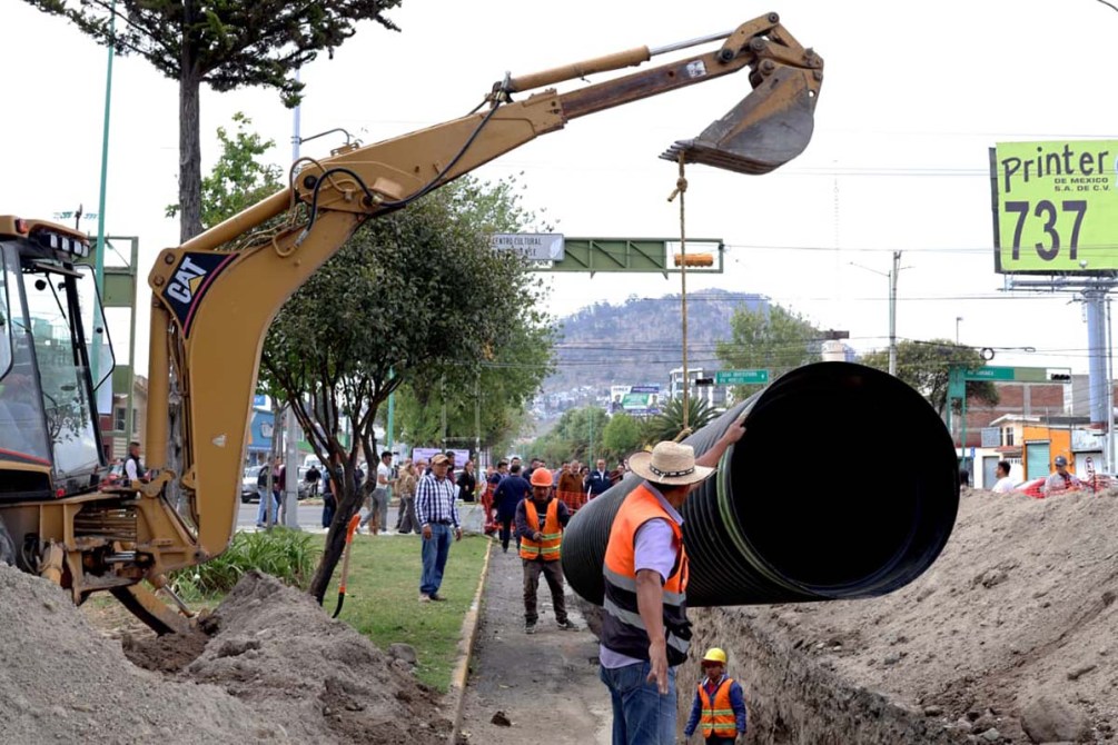 Construcción en una calle con una excavadora y trabajadores manejando una tubería grande.