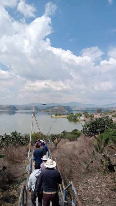 Grupo de personas caminando por un sendero hacia un lago, con montañas y nubes al fondo.