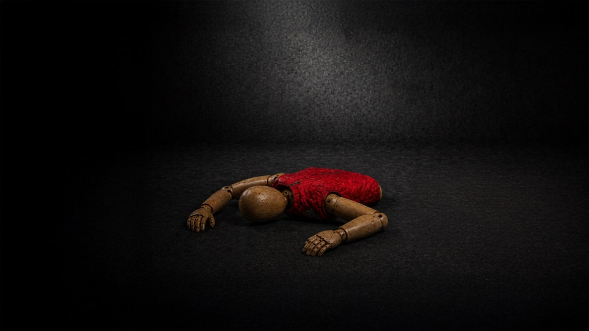 Figura de madera con un suéter rojo, tendida sobre una superficie oscura.
