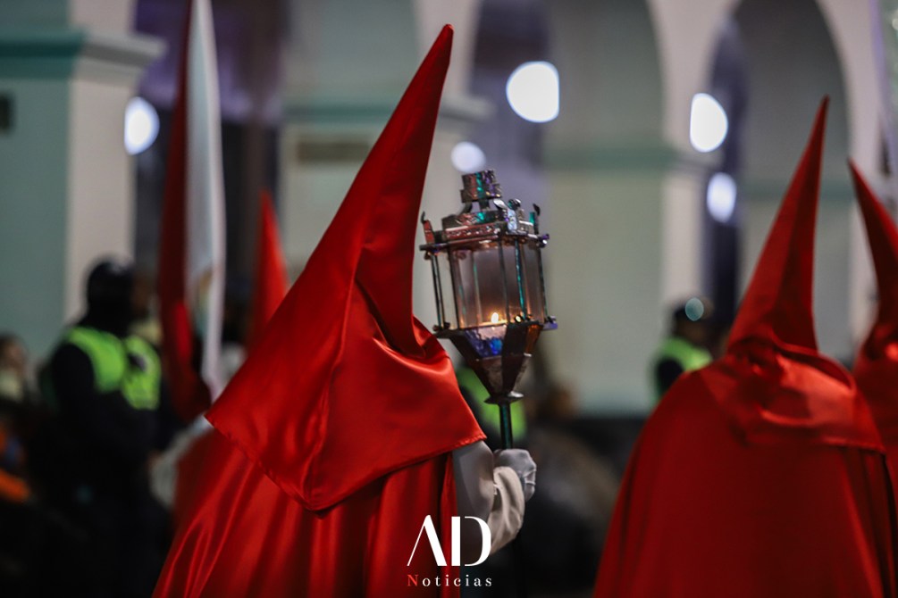 Personas vestidas con túnicas rojas y capirotes participan en una procesión, sosteniendo una linterna en un entorno nocturno.