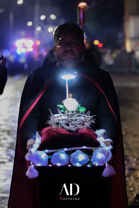 Hombre en una capa negra sosteniendo una ofrenda con luces y flores blancas en un ambiente nocturno.
